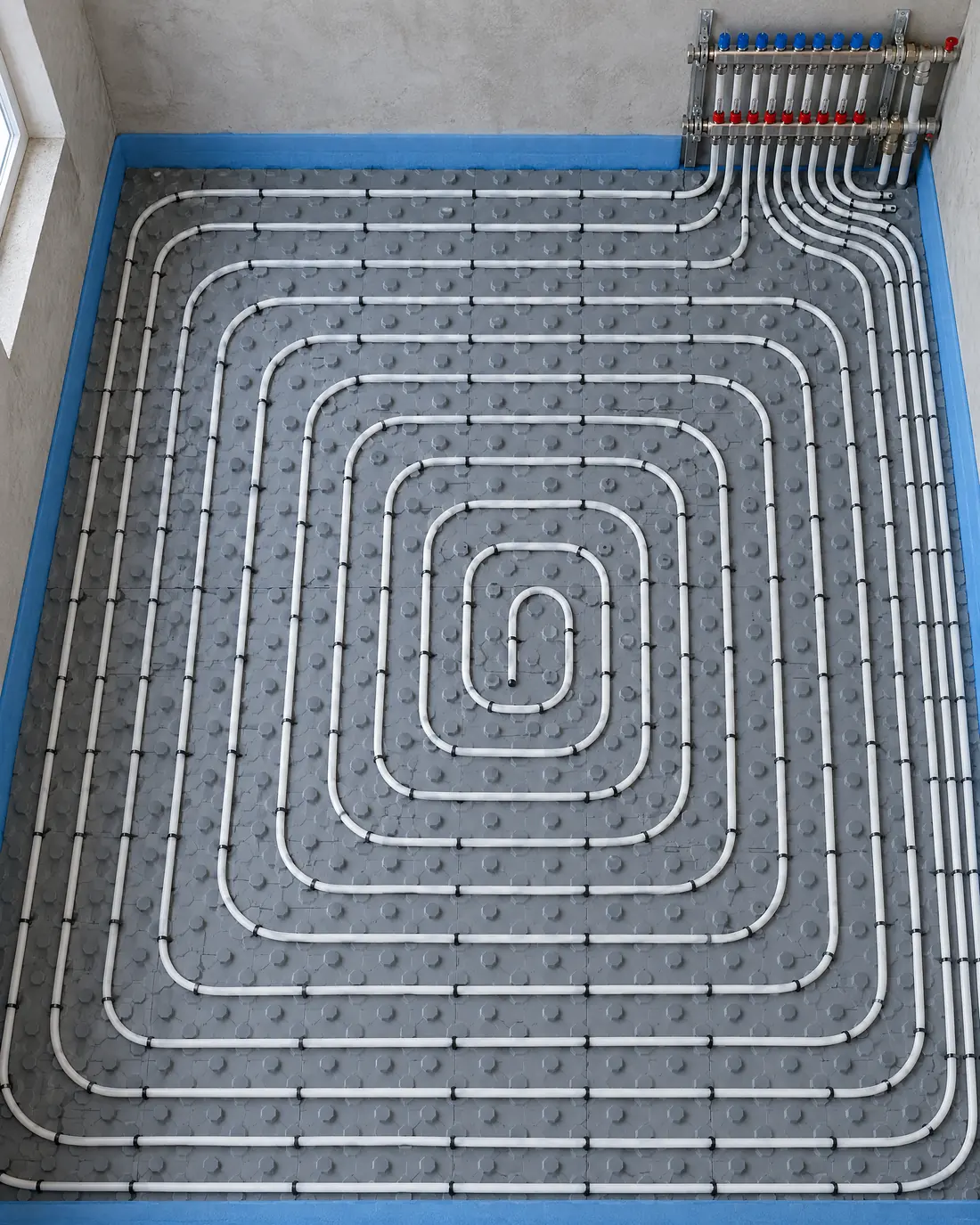 White heating pipes laid in a double spiral on insulation panels, with a manifold in the corner